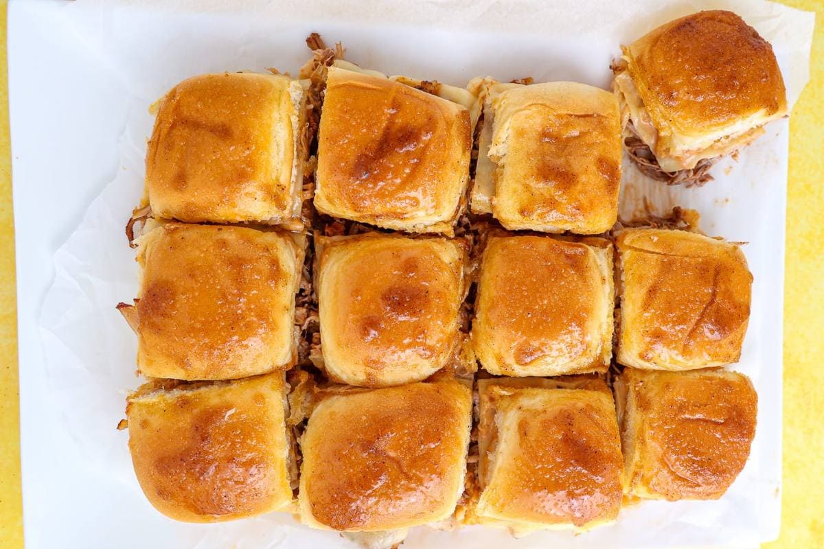 A tray of twelve golden-brown slider sandwiches, arranged in a grid on parchment paper, with one slider slightly separated from the rest.
