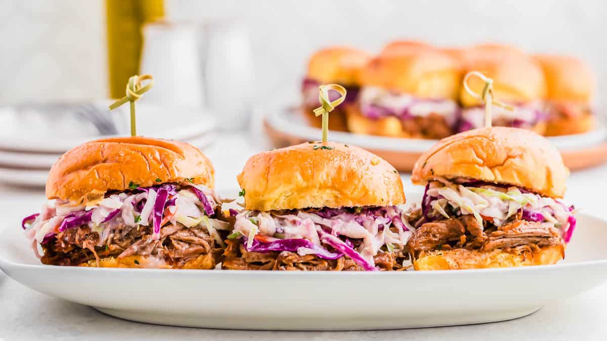 Three pulled pork sandwiches topped with coleslaw are arranged on a white plate, with more sandwiches blurred in the background.