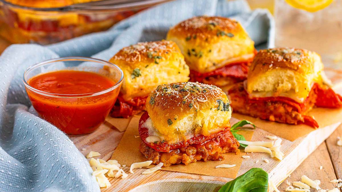 Four mini pizza sliders with pepperoni and cheese are arranged on parchment paper next to a small bowl of marinara sauce and sprinkled shredded cheese on a wooden board.