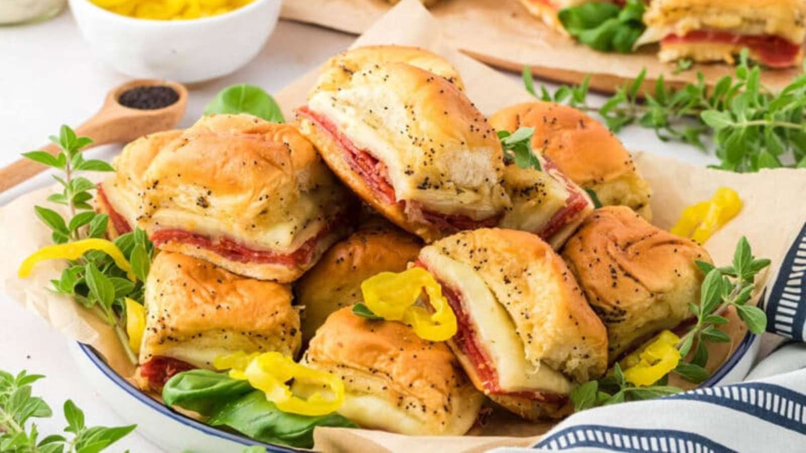 A plate of stacked baked sandwiches with layers of meat and cheese on soft buns, garnished with herbs and sliced yellow peppers.