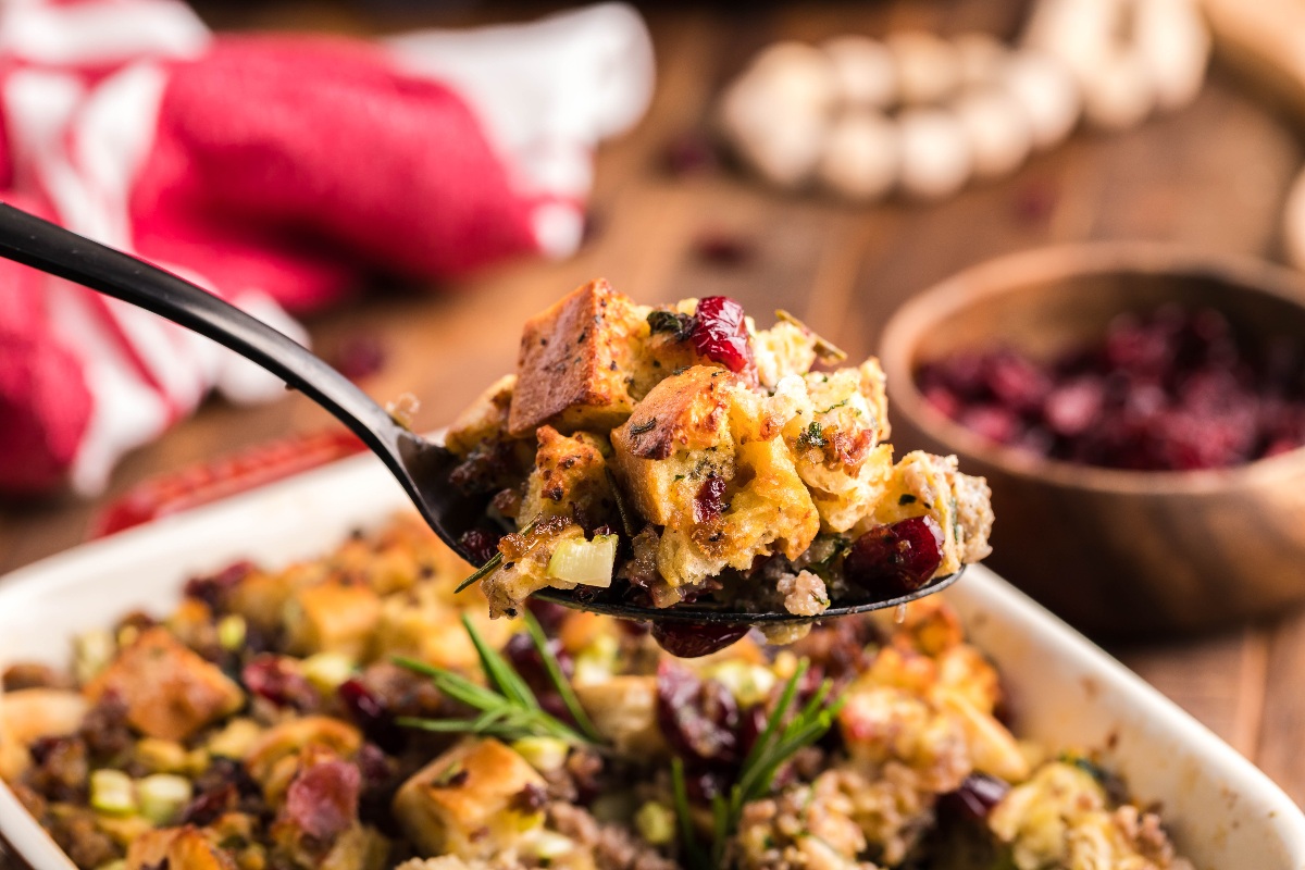 A spoon holds a serving of bread stuffing with herbs, cranberries, and visible chunks of vegetables above a casserole dish.