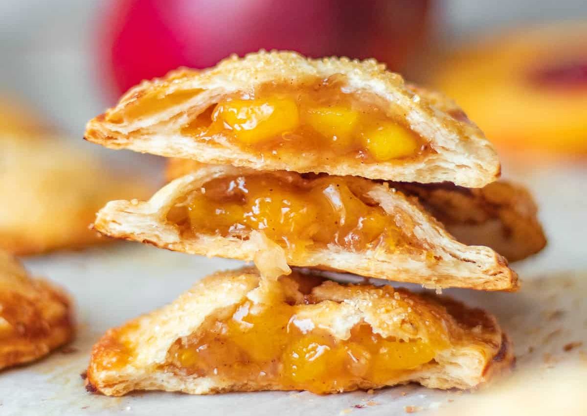 Three peach hand pies are stacked, each cut in half to reveal a golden, flaky crust and peach filling.