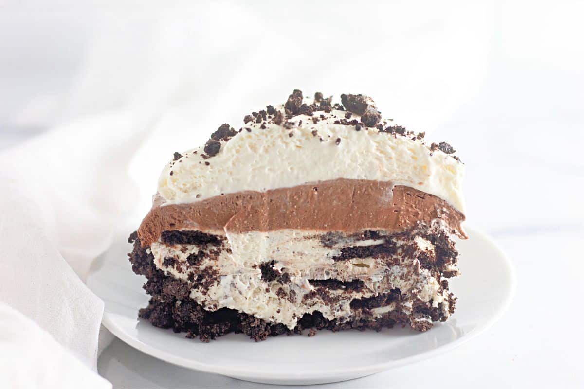 A slice of layered chocolate and cream dessert with a cookie crumb crust, topped with whipped cream and cookie crumbles, served on a white plate.
