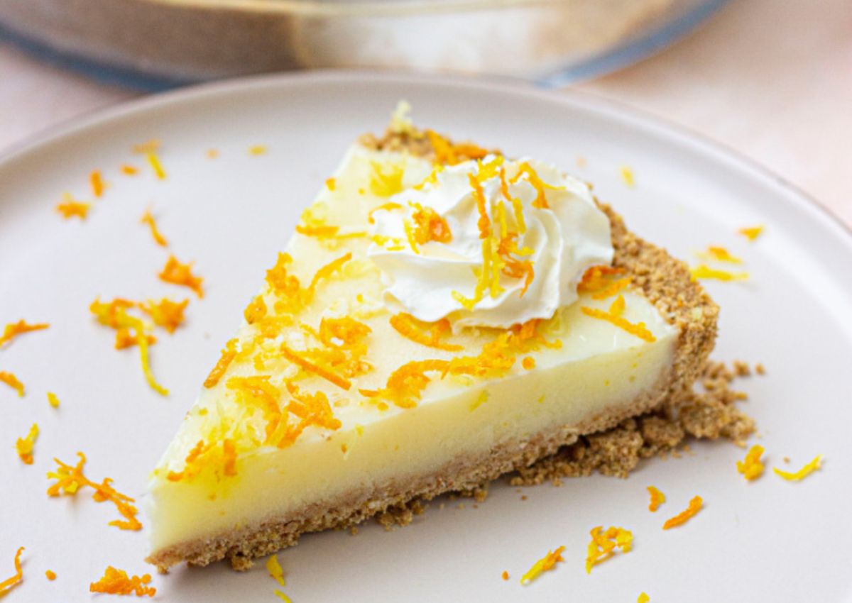 A slice of creamy pie with a graham cracker crust, topped with whipped cream and orange zest, on a white plate.