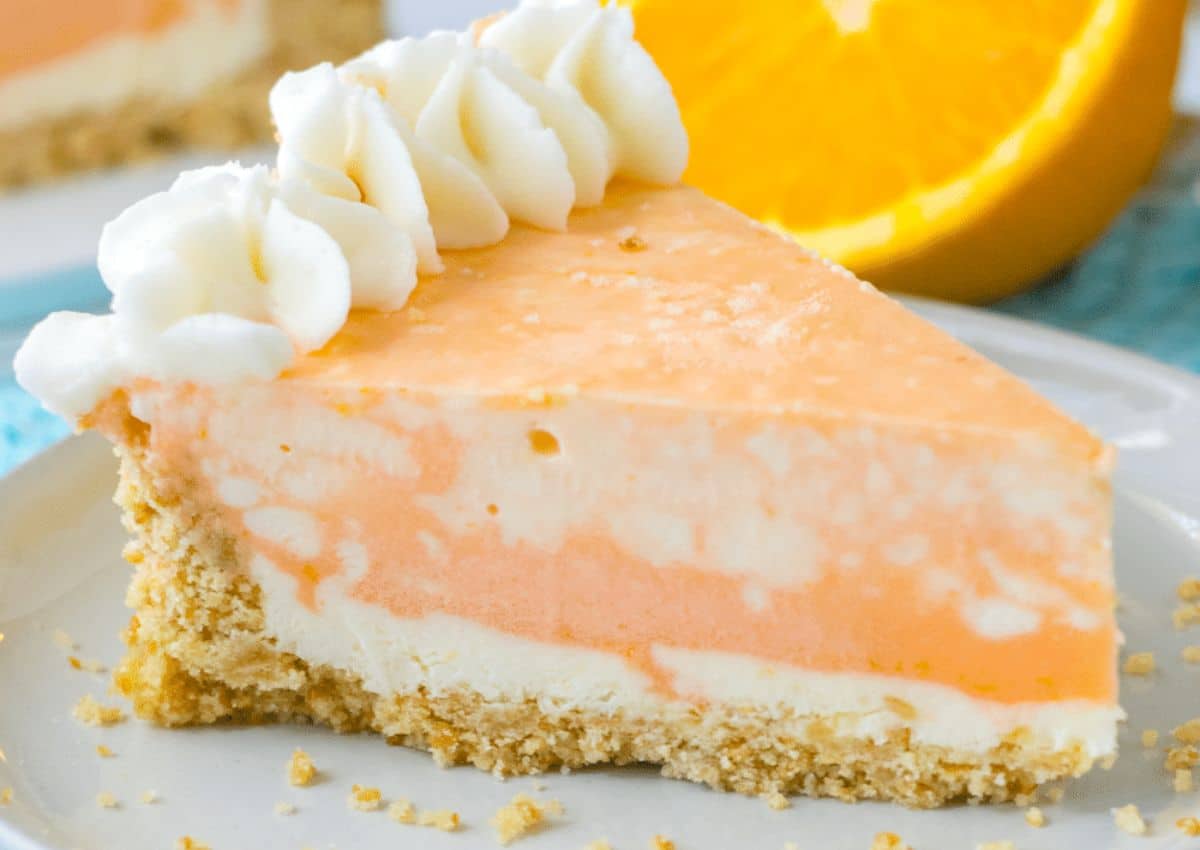 A slice of creamy orange and white cheesecake with a crumbly crust, topped with whipped cream, sits on a plate with an orange in the background.
