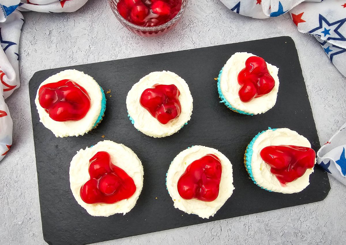 Six cupcakes with white frosting and cherry topping are arranged on a black rectangular slate. A bowl of extra cherry topping and a star-patterned cloth are nearby.