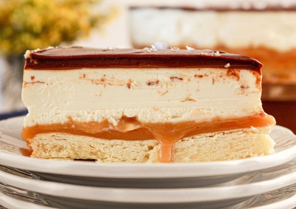 A close-up of a dessert slice with layers of shortbread, caramel, creamy filling, and chocolate glaze, served on a white plate.