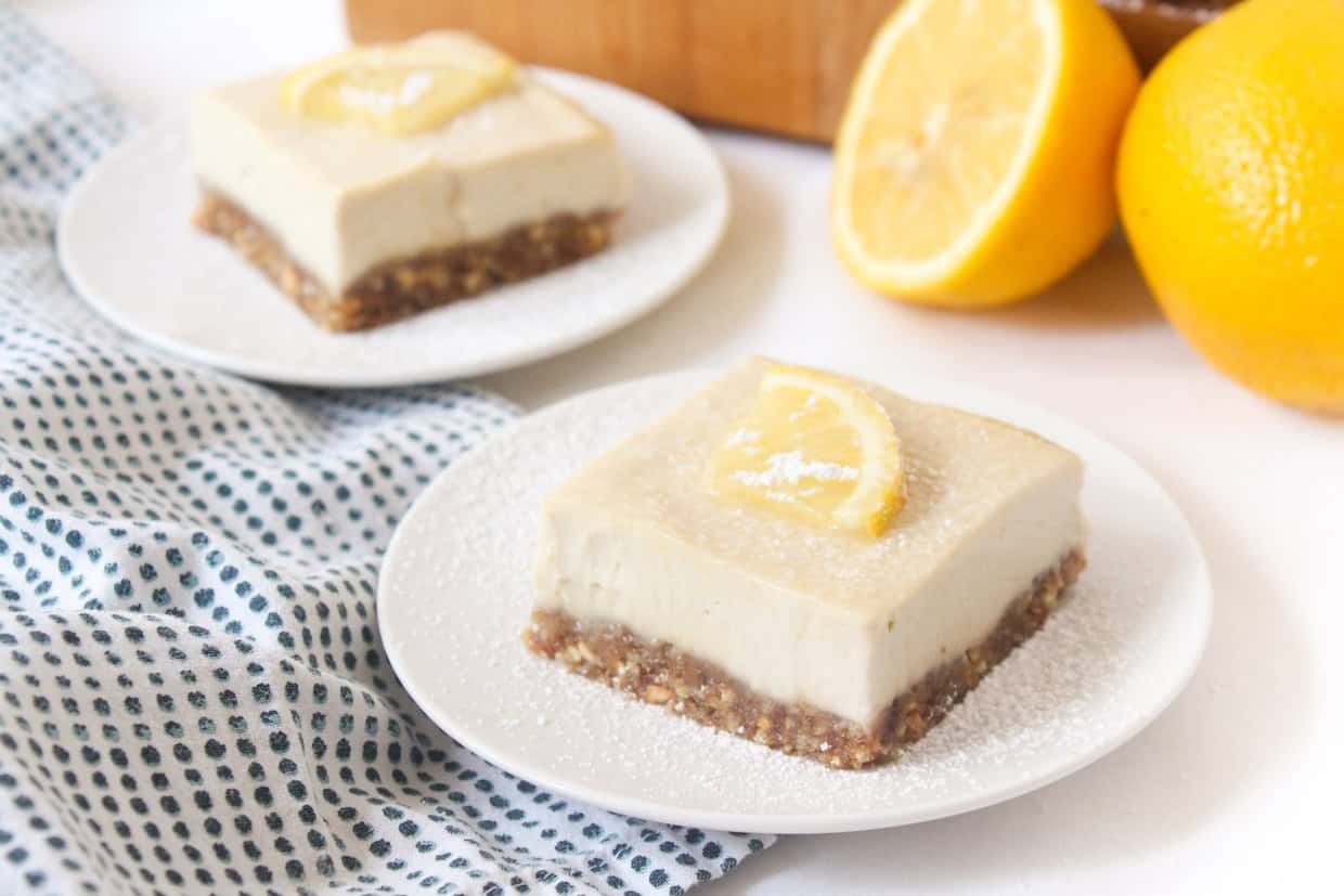 Two lemon bars with a nutty crust and creamy topping, garnished with a lemon slice, served on white plates beside a blue dotted cloth and fresh lemons.