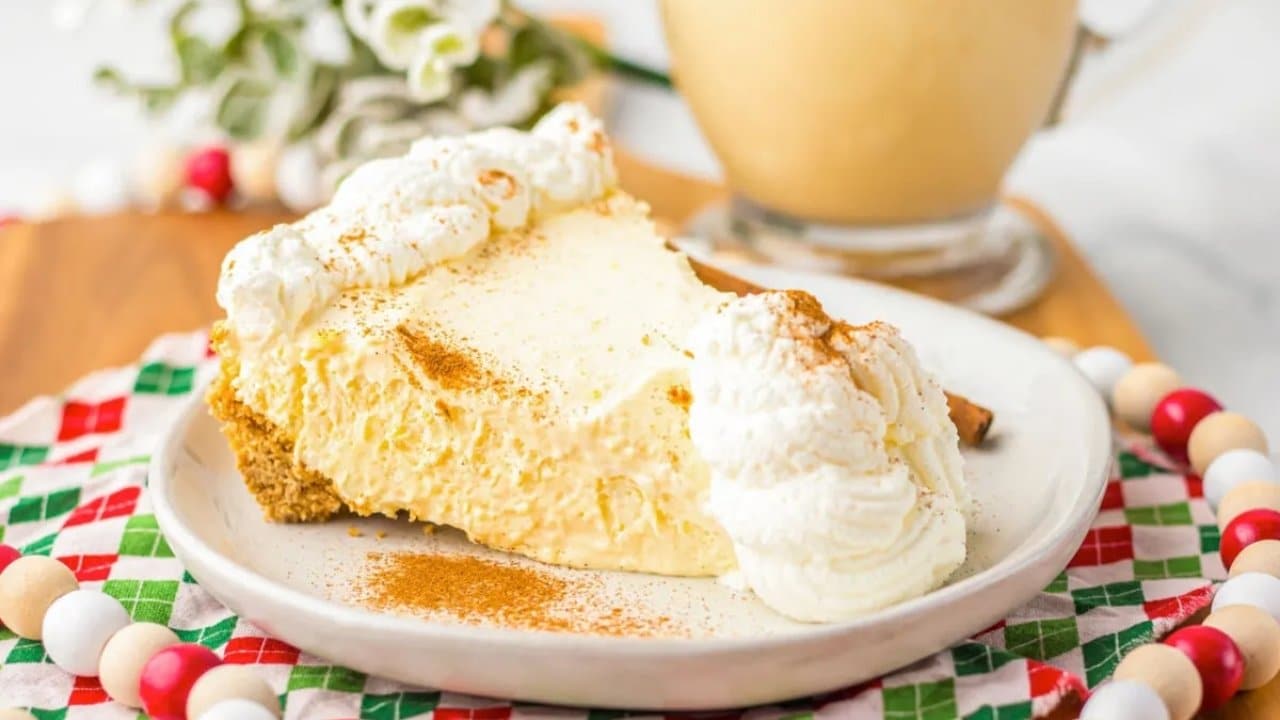 A slice of creamy pie with whipped cream and cinnamon on top sits on a white plate, with a festive napkin and a glass of eggnog in the background.