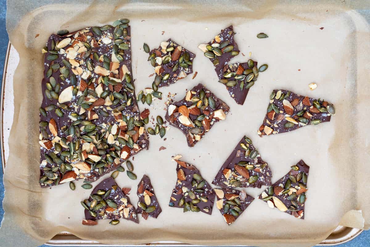 Broken pieces of dark chocolate bark topped with pumpkin seeds and chopped nuts on a parchment-lined baking tray.