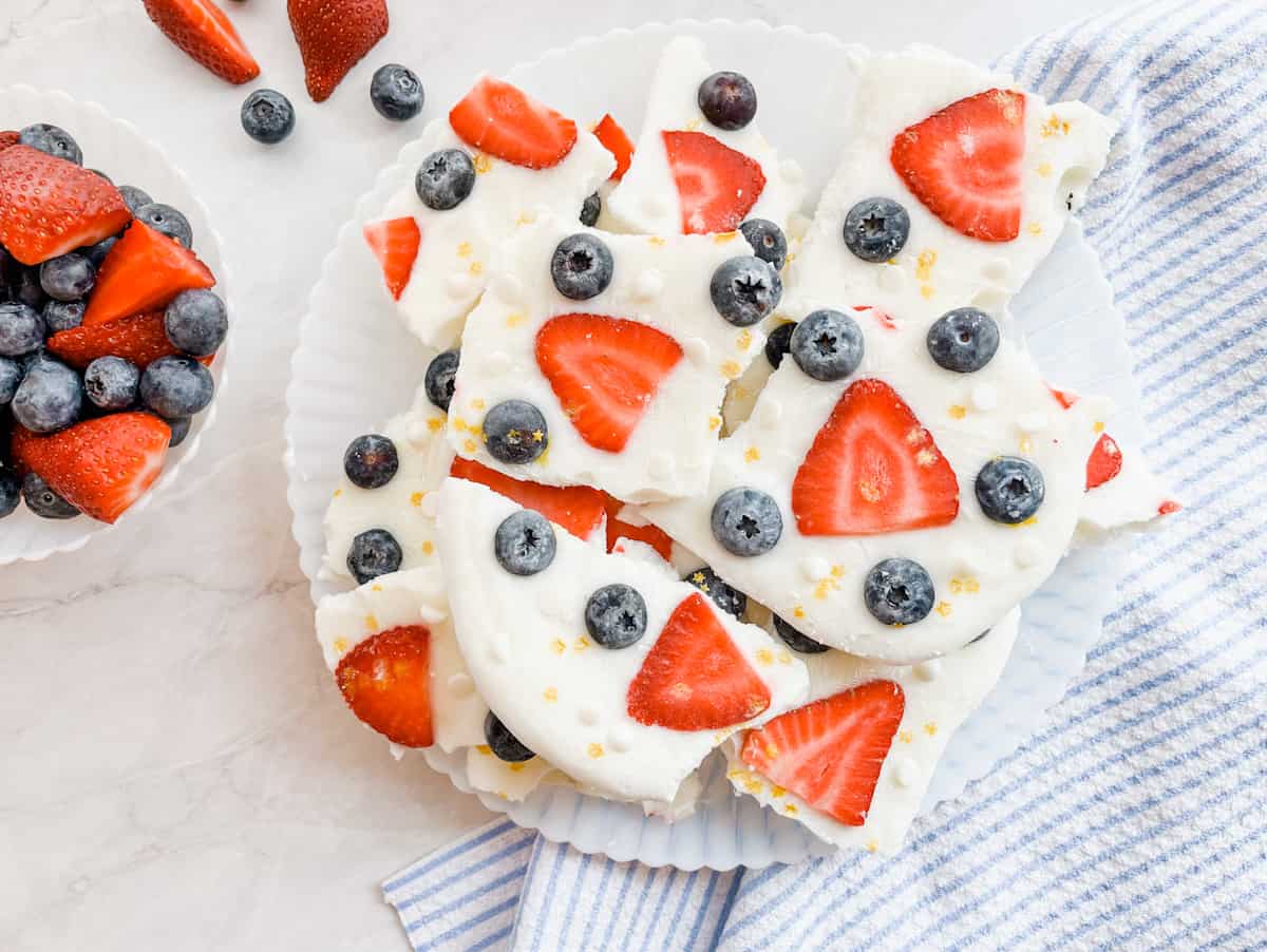 A plate of yogurt bark pieces topped with sliced strawberries and whole blueberries, next to a bowl of fresh berries and a striped cloth.