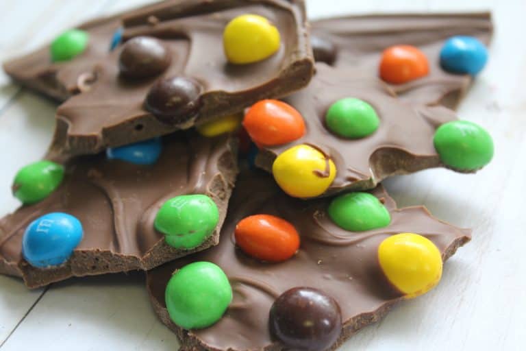 Pieces of chocolate bark topped with colorful candy-coated chocolates are stacked on a white surface.