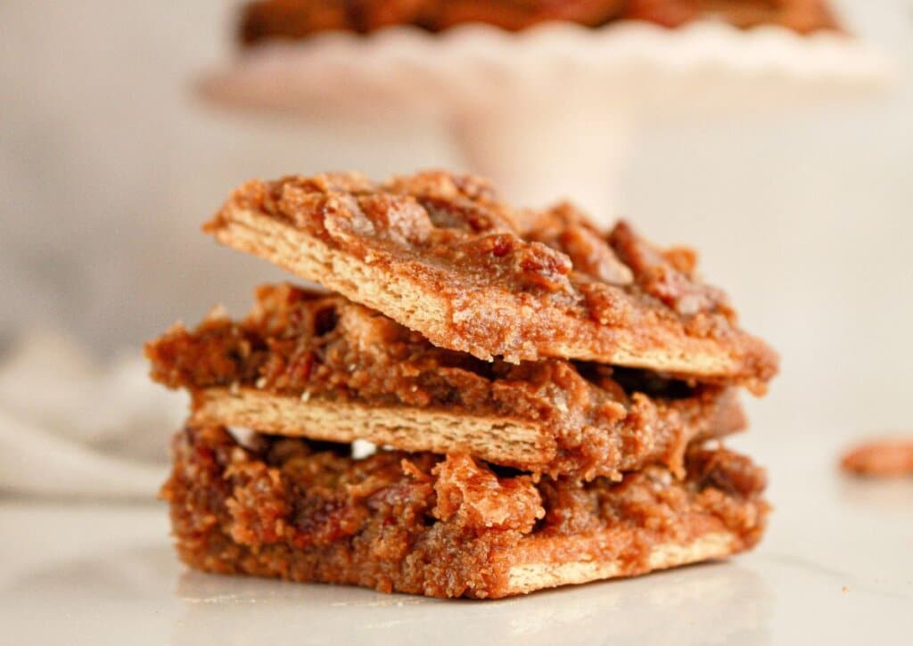 Three pecan pie bars are stacked on a white surface, showing a crumbly nut topping and a crisp crust.
