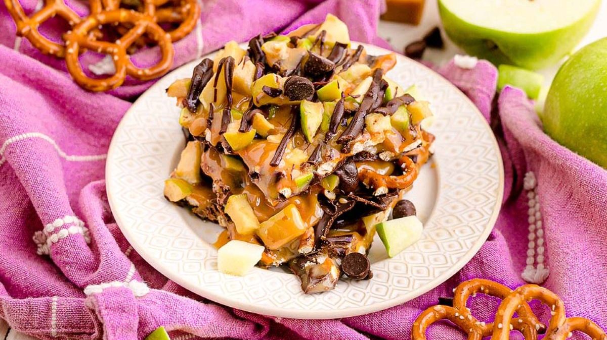 A plate of caramel apple bark with pretzels, apple pieces, chocolate chips, and caramel drizzle, served on a white plate over a purple cloth.
