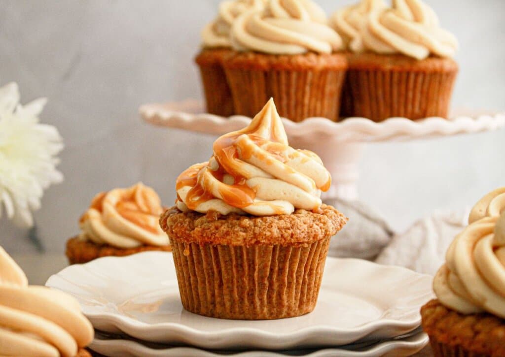 A cupcake with swirled caramel frosting sits on stacked white plates, with more cupcakes displayed in the background.