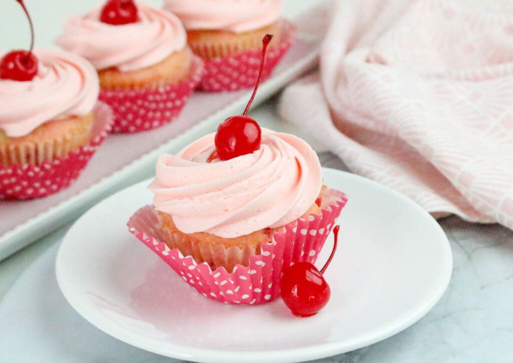 A cherry-topped cupcake with pink frosting in a pink wrapper on a white plate, with two cherries beside it and more cupcakes in the background.