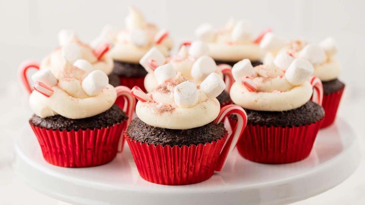Six chocolate cupcakes in red wrappers topped with white frosting, mini marshmallows, crushed candy canes, and small candy cane handles displayed on a white cake stand.