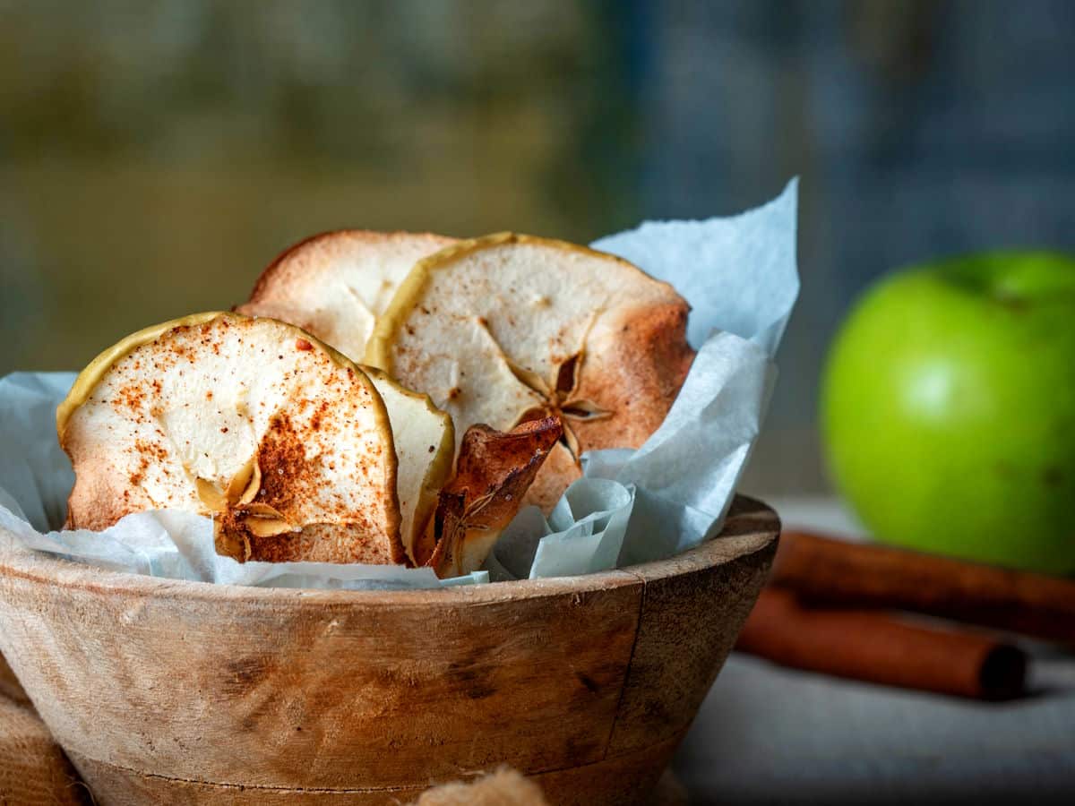 Wooden bowl with dried apple slices on parchment paper, a green apple, and cinnamon sticks in the background.