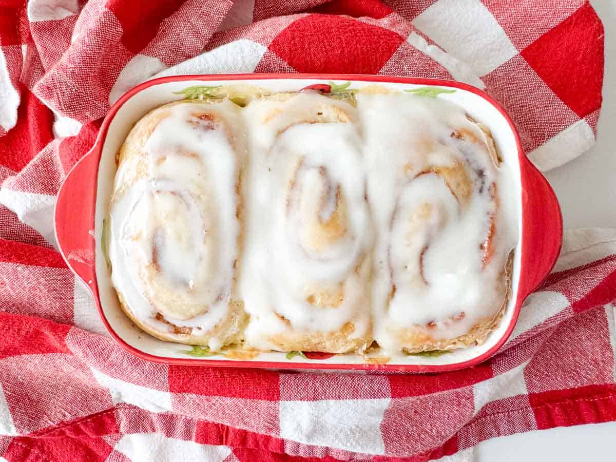 Three frosted cinnamon rolls in a red baking dish, placed on a red and white checkered cloth.