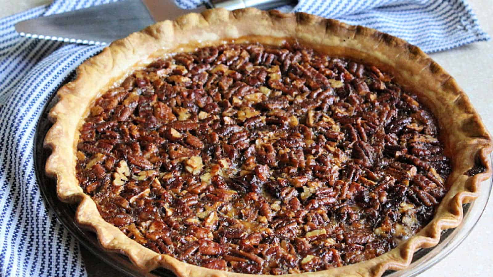 A baked pecan pie with a golden, crimped crust sits on a striped cloth next to a metal pie server.