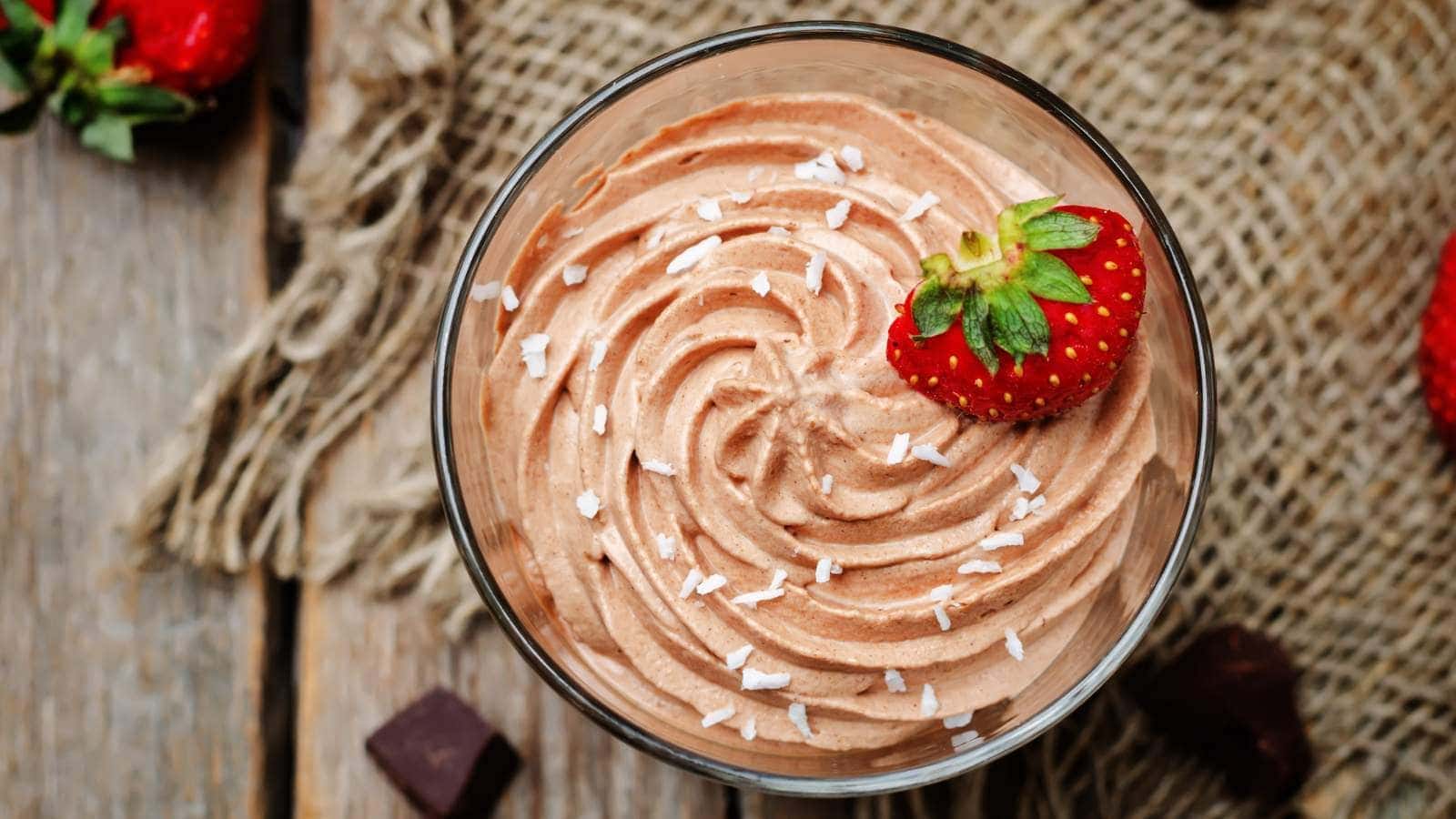 A glass of chocolate mousse topped with shredded coconut and a whole strawberry, placed on a wooden table with pieces of chocolate and burlap fabric in the background.