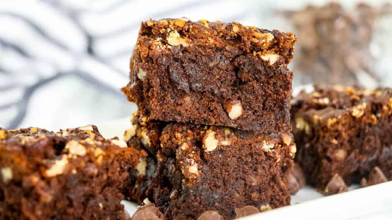 Close-up of three chocolate brownies with visible nuts stacked on a white surface, with more brownies and chocolate chips in the background.