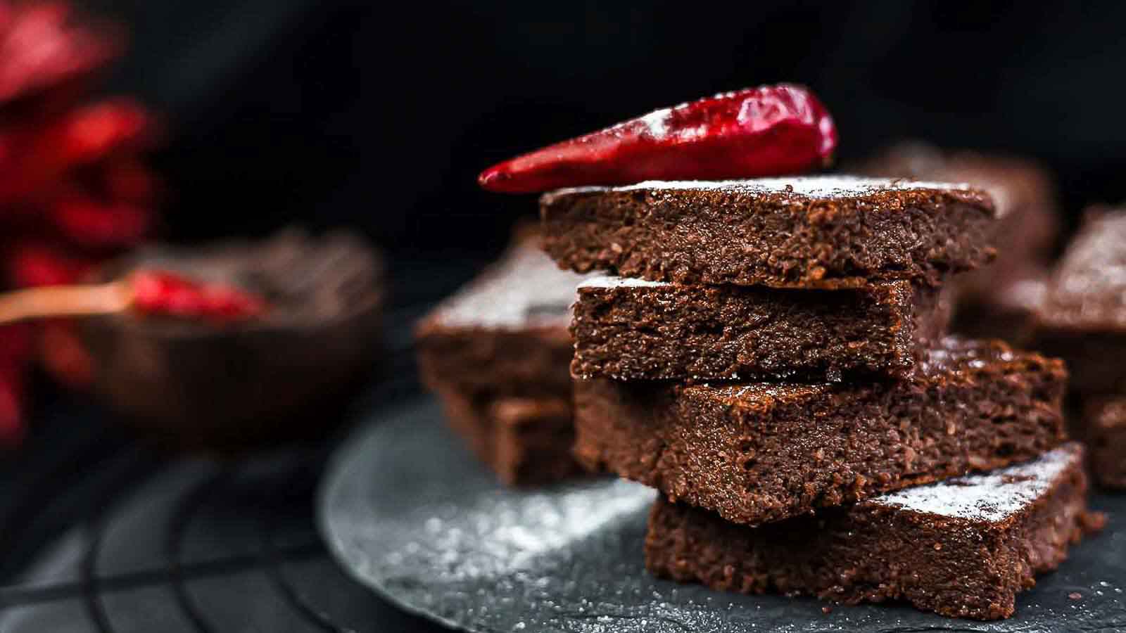 A stack of chocolate brownies topped with a red chili pepper sits on a dark plate, with more brownies and a blurred red object in the background.