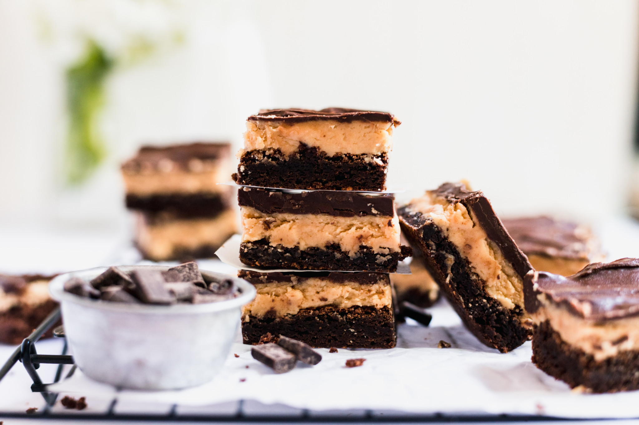Three chocolate and peanut butter layered dessert bars are stacked on a wire rack, with more bars and a small metal cup of chocolate chunks nearby.