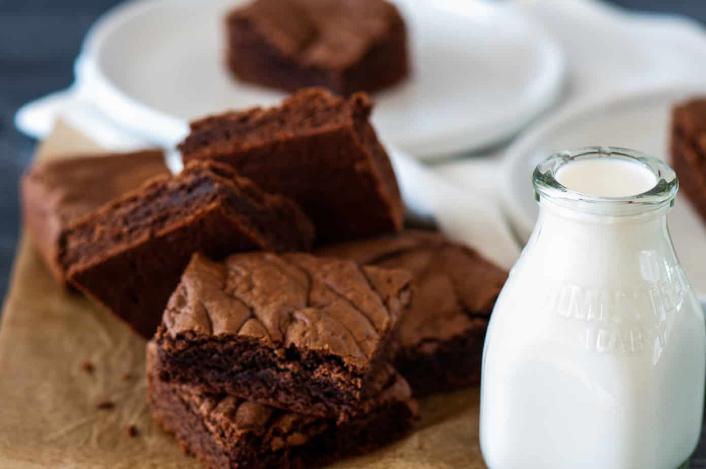 Several chocolate brownies are stacked on parchment paper next to a small glass bottle of milk, with a white plate and napkin in the background.