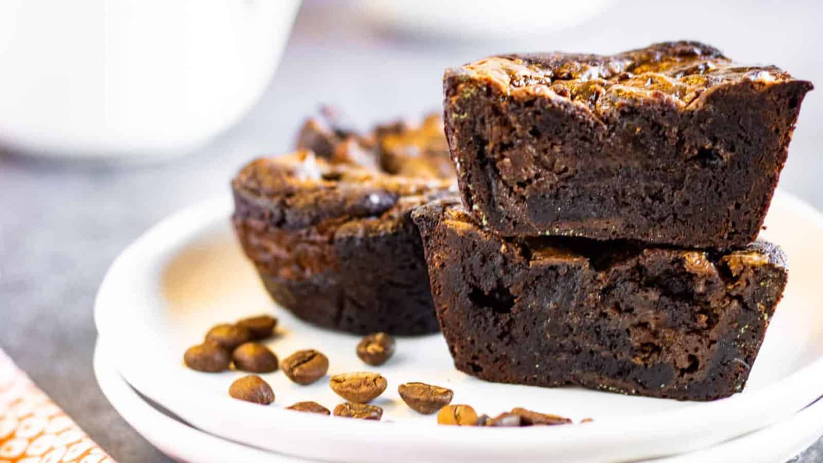 Two chocolate brownies are stacked on a white plate with coffee beans scattered around them.