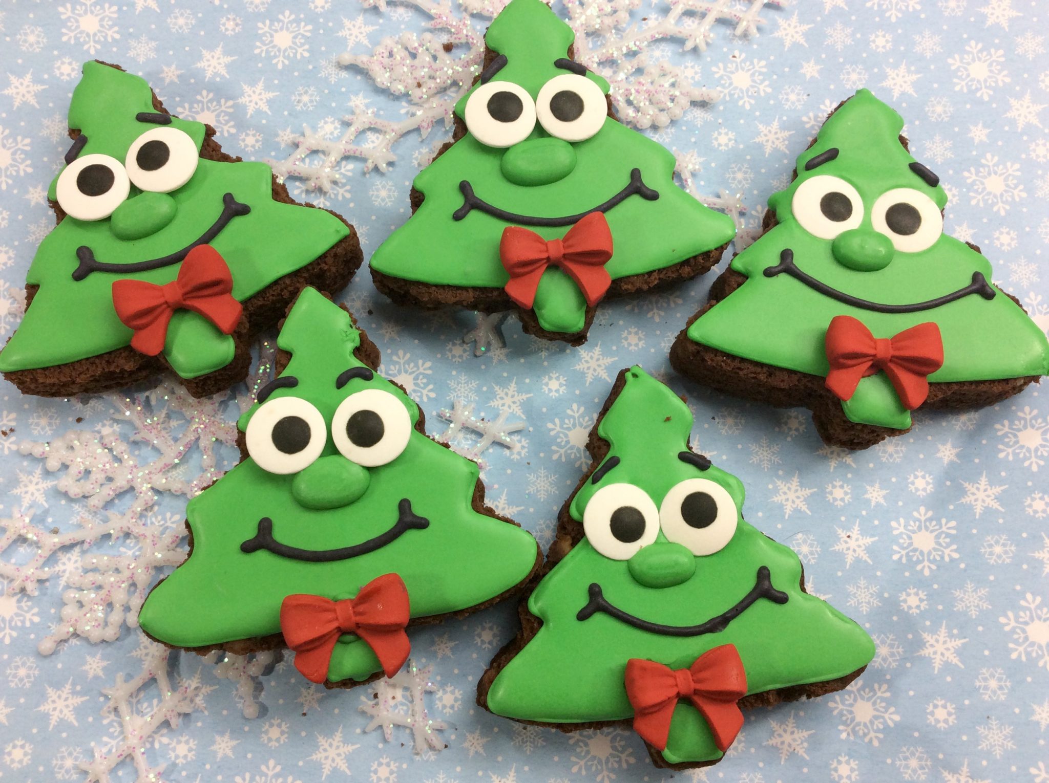 Five Christmas tree-shaped cookies decorated with green icing, smiling faces, red bow ties, and placed on a snowflake-patterned background.