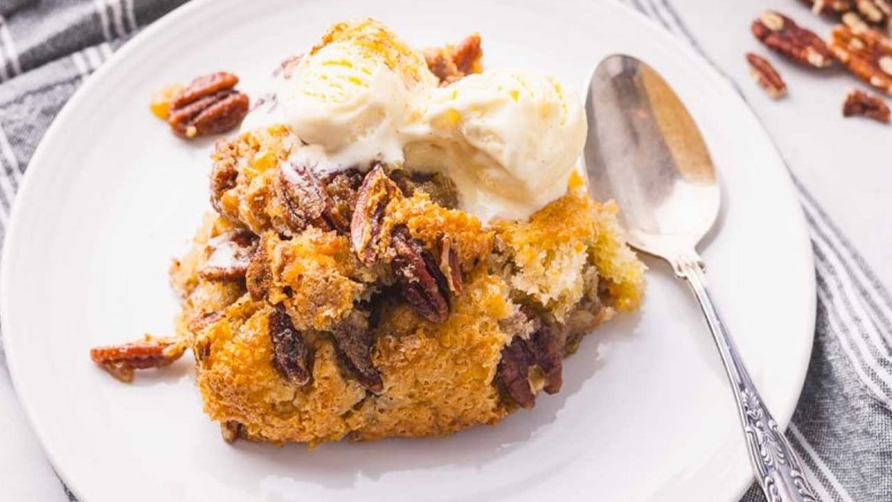 A serving of pecan cobbler topped with vanilla ice cream is on a white plate with a spoon beside it.