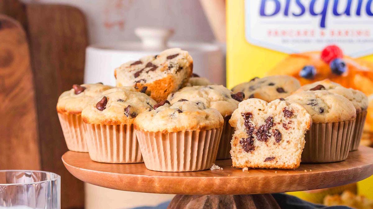 A wooden cake stand displays chocolate chip muffins, one of which is cut in half to show the inside; a Bisquick box is visible in the background.