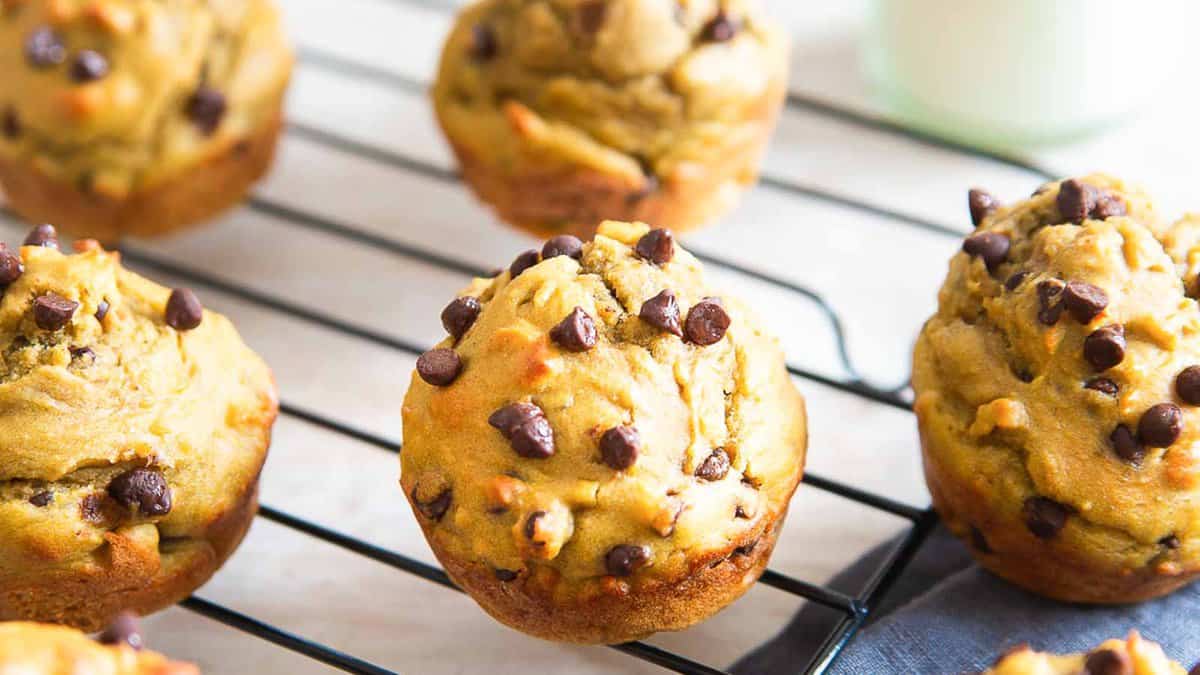 Freshly baked chocolate chip muffins are cooling on a wire rack, with a few more muffins and a glass of milk visible in the background.