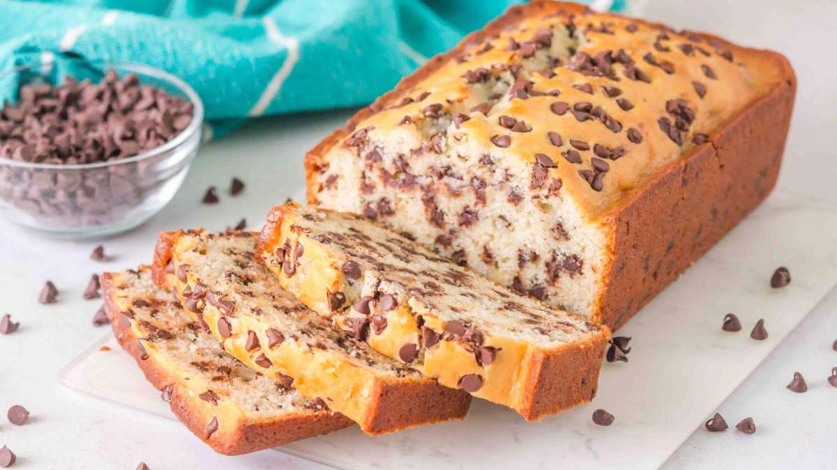 A loaf of chocolate chip bread is sliced on a white cutting board, with a bowl of chocolate chips and a teal cloth in the background.