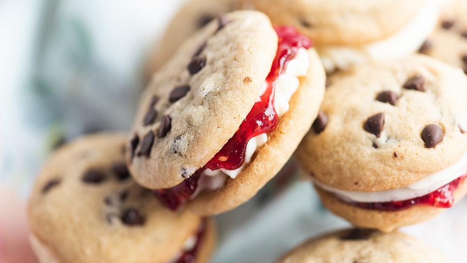A close-up of chocolate chip cookie sandwiches filled with white cream and strawberry jam.