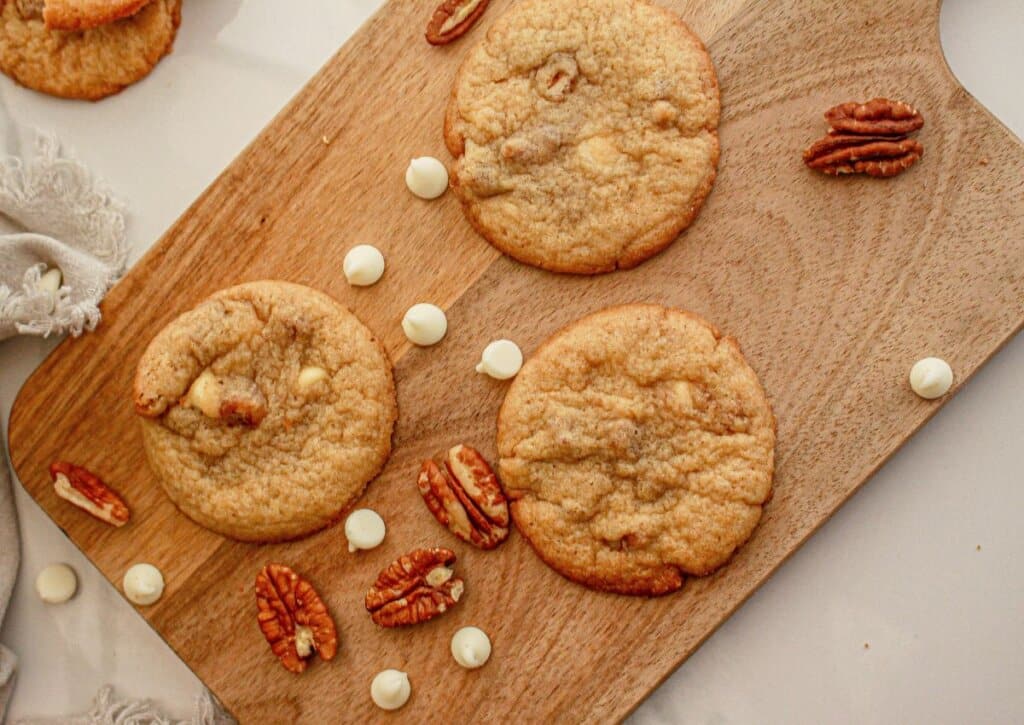 Three cookies are placed on a wooden cutting board, surrounded by pecan halves and white chocolate chips.