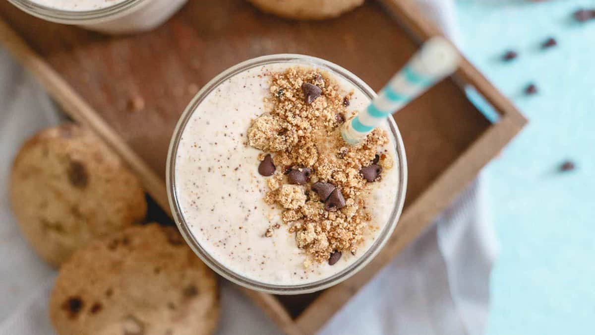 A glass of creamy milkshake topped with crumbled cookies and chocolate chips, with a striped straw, placed on a tray next to cookies.