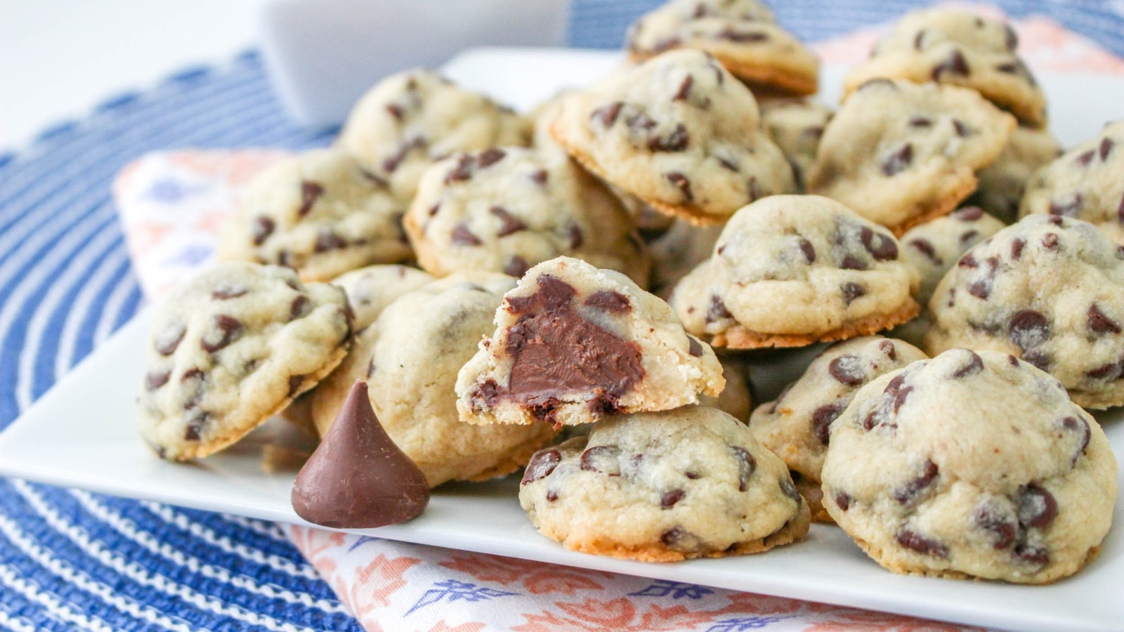 A plate of chocolate chip cookies, some with a chocolate filling visible inside, sits on a patterned cloth with a chocolate candy in the foreground.