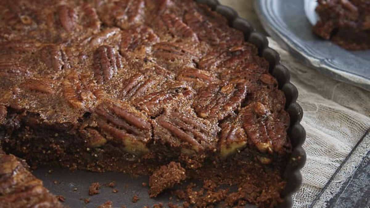 A close-up of a pecan pie with a slice removed, showing its nut topping and rich filling inside a fluted tart pan.