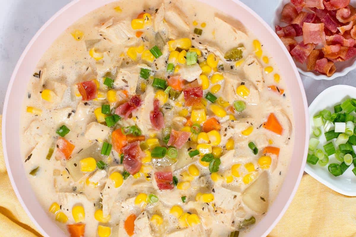 A bowl of creamy chicken and corn chowder topped with chopped green onions and bacon bits, with additional toppings in small bowls on the side.