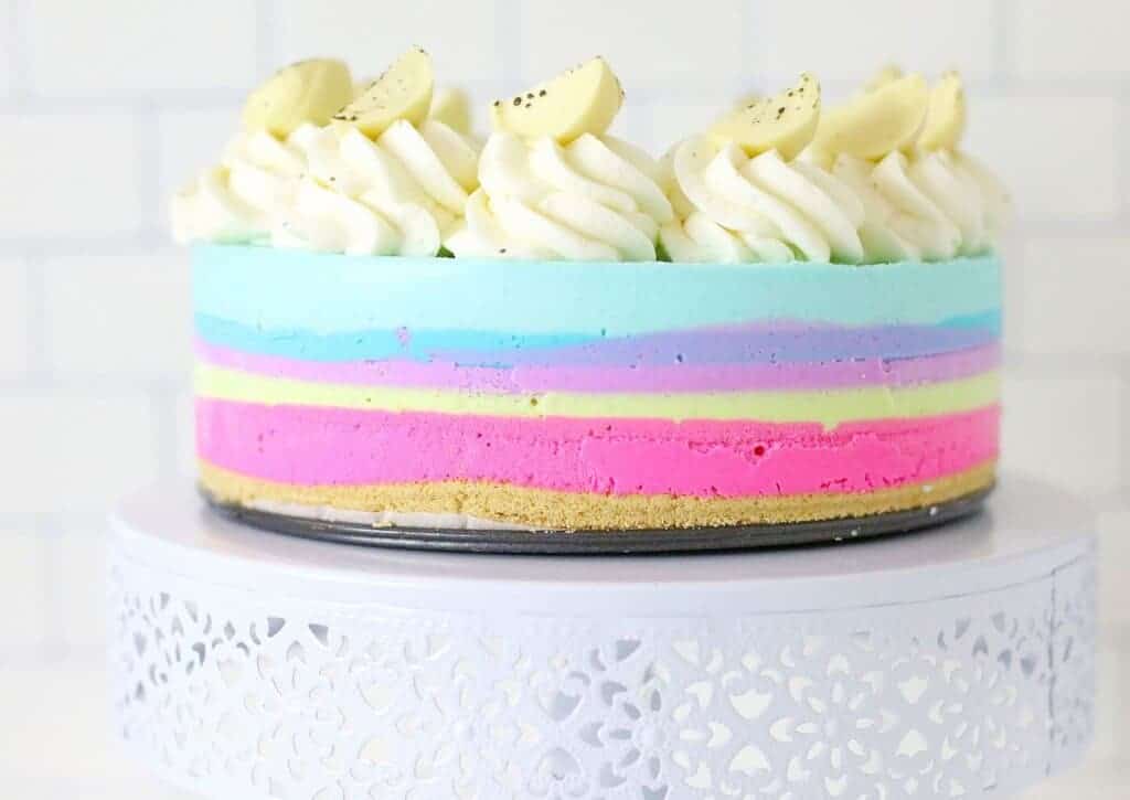A pastel-colored, multi-layered cheesecake with a graham cracker crust, topped with piped white cream and white chocolate pieces, displayed on a white decorative cake stand.