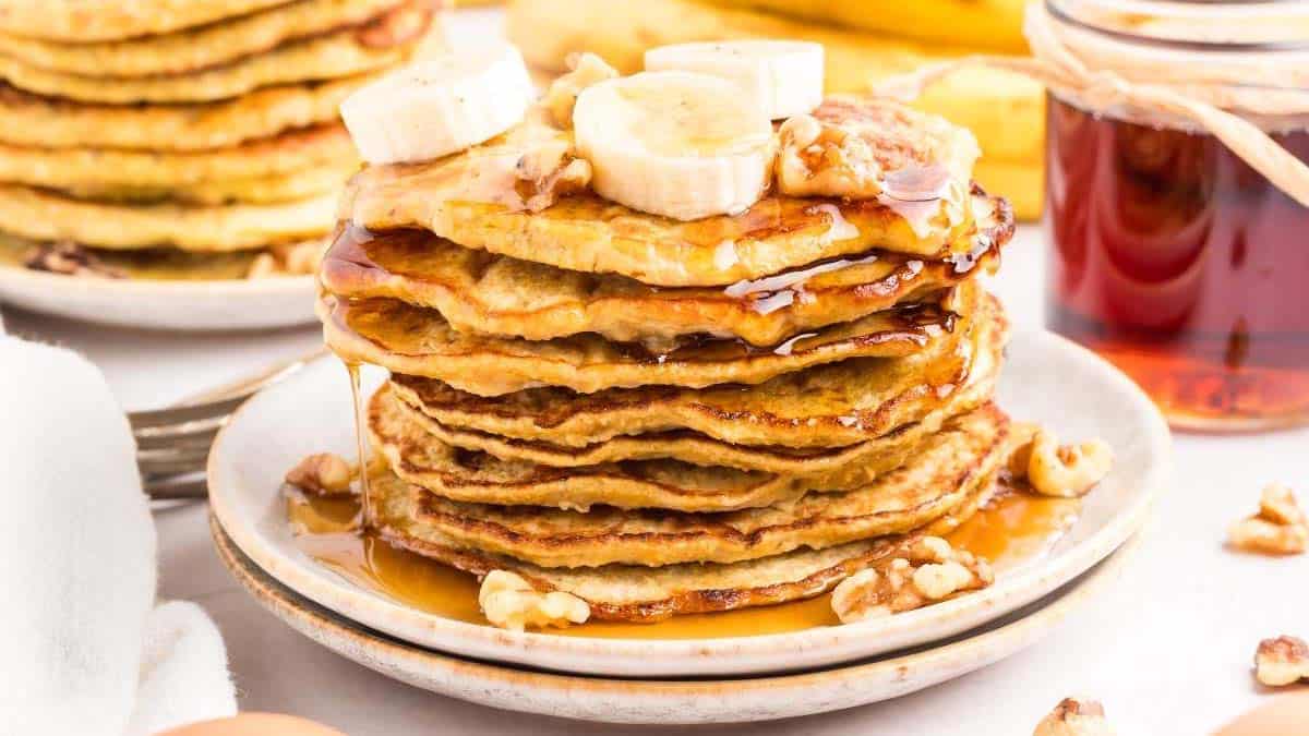 A stack of pancakes topped with banana slices, chopped nuts, and syrup sits on a white plate, with a jar of syrup and more pancakes in the background.