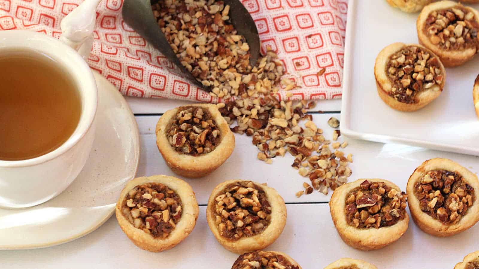 A cup of tea sits beside a group of mini pecan tarts, with chopped nuts spilling from a scoop onto a white surface near a red-patterned cloth.