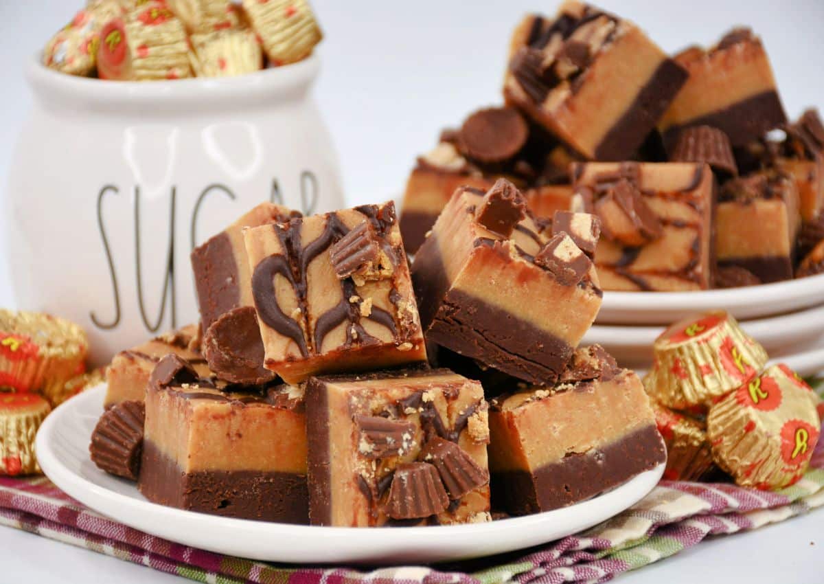 Assorted chocolate and peanut butter fudge squares stacked on plates, surrounded by mini peanut butter cups and a jar labeled SUGAR in the background.