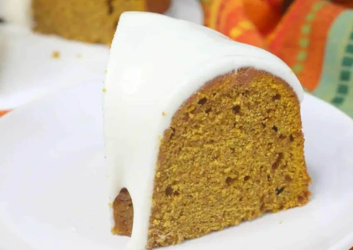 A slice of bundt cake with orange-brown crumb and white icing sits on a white plate.
