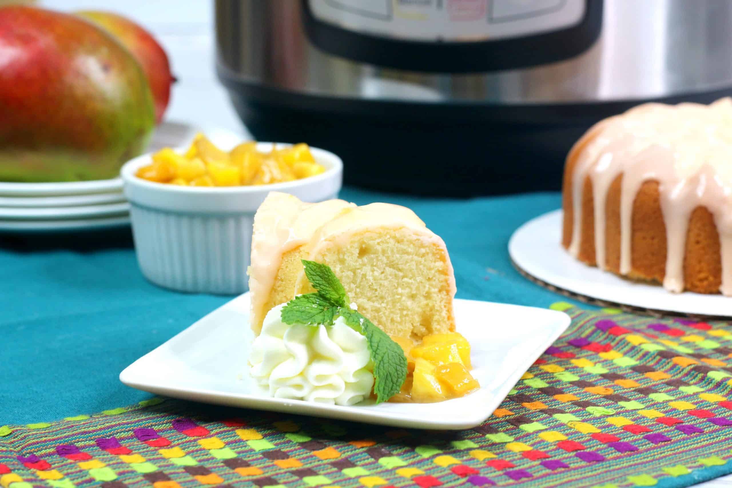 A slice of bundt cake with whipped cream, mango pieces, and a mint leaf on a white plate, with a whole bundt cake, mangoes, and a kitchen appliance in the background.