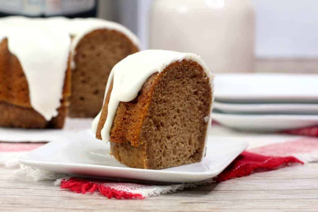 A slice of bundt cake with white icing is served on a white plate, with the rest of the cake and stacked plates in the background.