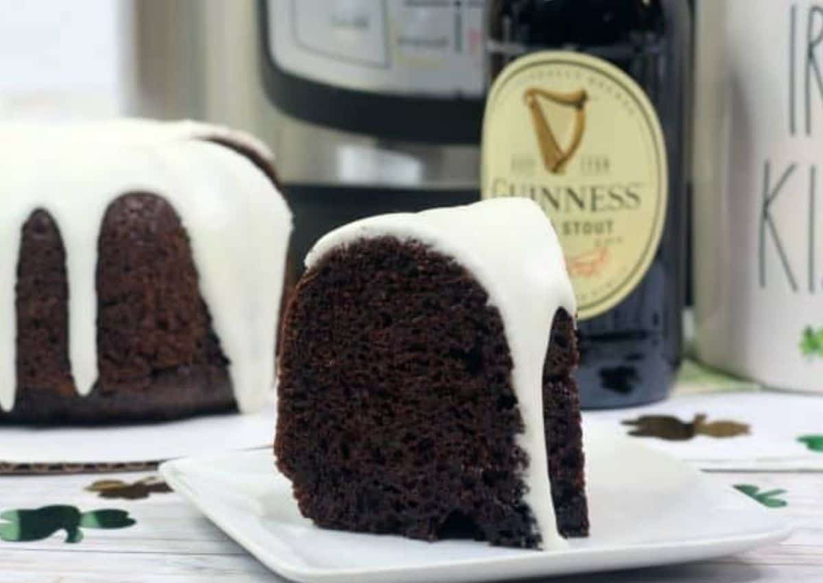 A slice of chocolate bundt cake with white icing sits on a plate, with the rest of the cake and a bottle of Guinness stout in the background.