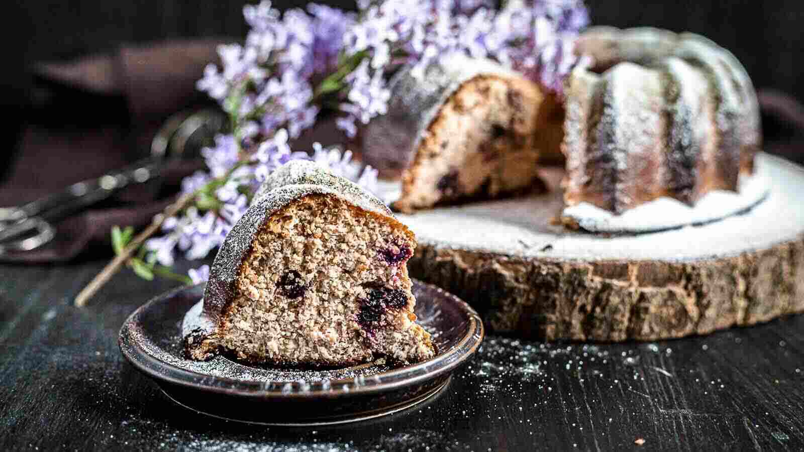 A slice of bundt cake with visible berries sits on a plate in front of the remaining cake, garnished with powdered sugar and purple flowers in the background.